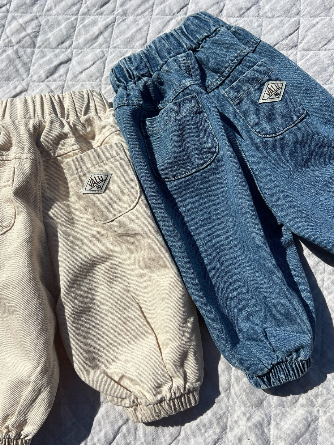 Denim | Two pairs of BALLOON JEAN pants with elastic waistbands and cuffs rest on a quilted surface. One beige, the other blue, each has a logo patch above the back pocket. With their jogger-style fit, these Valu Baby essentials offer comfort and style for any young adventurer.