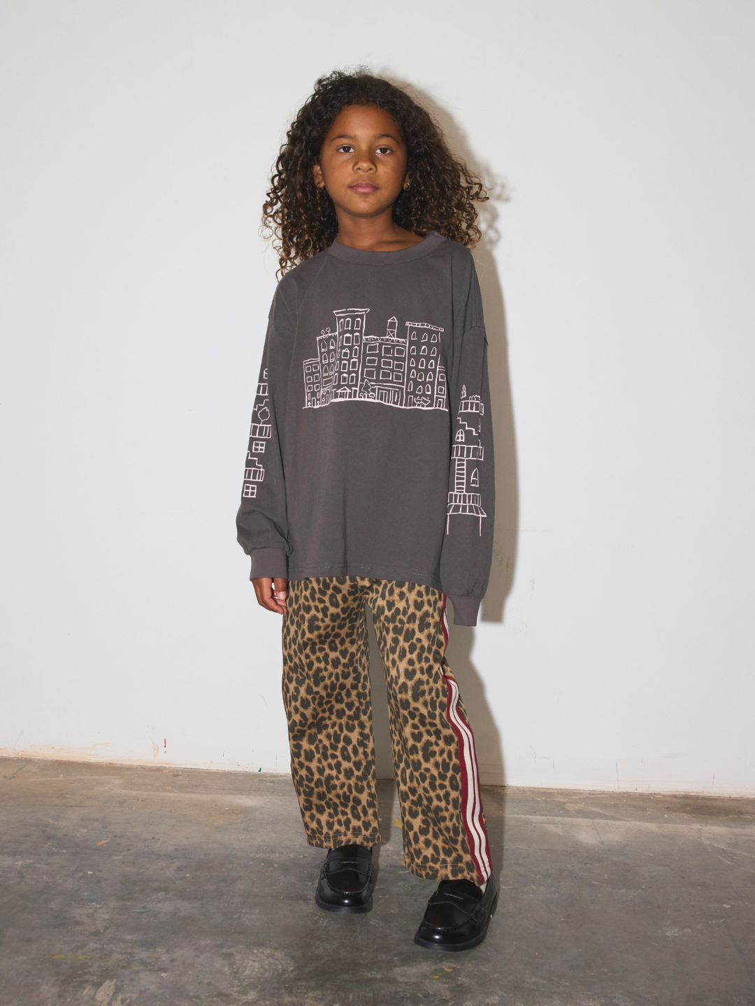 A child wearing a dark gray crewneck sweatshirt with a white graphic print on the front and sleeves, paired with leopard print pants featuring a red and white side stripe, and black shoes, standing against a plain white wall.