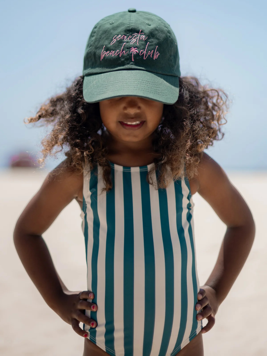 A young child in the CABANA STRIPE ONE PIECE SWIMSUIT and a green cap stands confidently on a sandy beach, hands on hips, with the ocean softly blurred in the background.