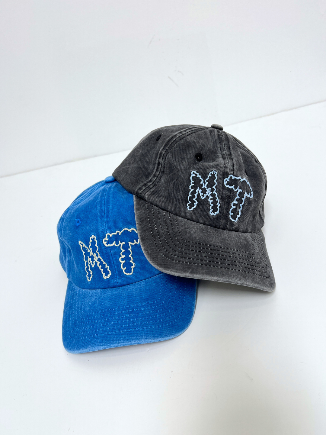 Blue | Two MT DAD CAPs in blue and black cotton canvas, both featuring "NT" (Milk Teeth) embroidered in white cloud-style font, overlap on a white surface.