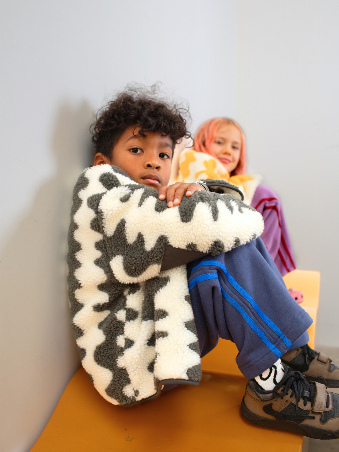 Child sitting on a chair with a patterned jacket, another child in the background.