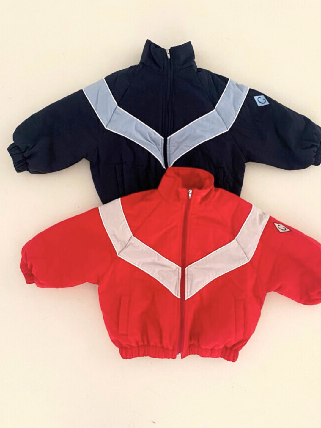 Navy | Two children's WARM-UP JACKETS are displayed. One, a retro track jacket in black with light blue and white chevron designs, and the other, red with white and gray chevron designs. Both from a popular Korean label, they feature zippers and elastic cuffs for a stylish, lightweight layer.