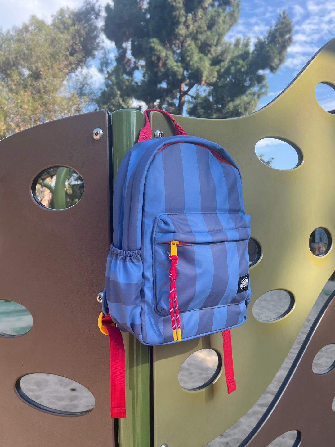 Blue Stripe | A blue and darker blue striped backpack with red straps and a red zipper pull hanging on a playground metal structure outdoors.