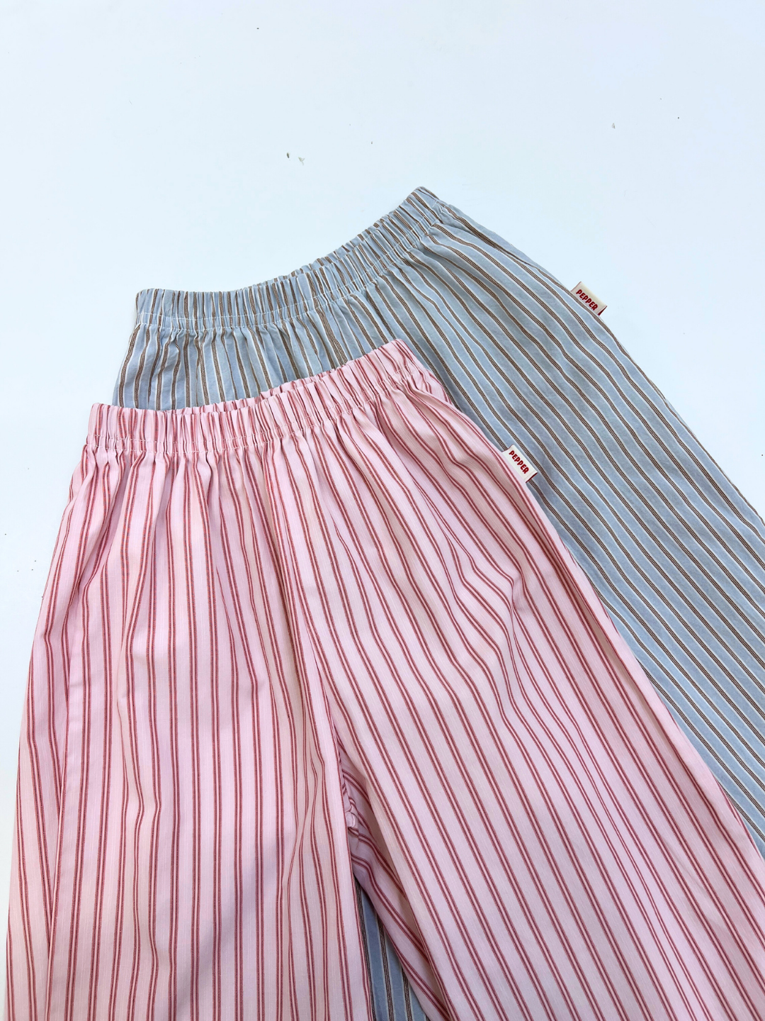 Pink | Two POPLIN STRIPE PANT pairs—one pink with red stripes, one light blue with gray stripes—are neatly laid flat on a white surface.