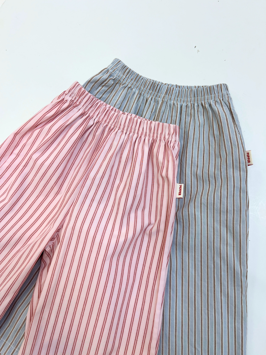 Two POPLIN STRIPE PANT pairs are displayed: one in pink with red stripes, the other in gray with white stripes. Both feature elastic waists and are neatly arranged on a white surface.