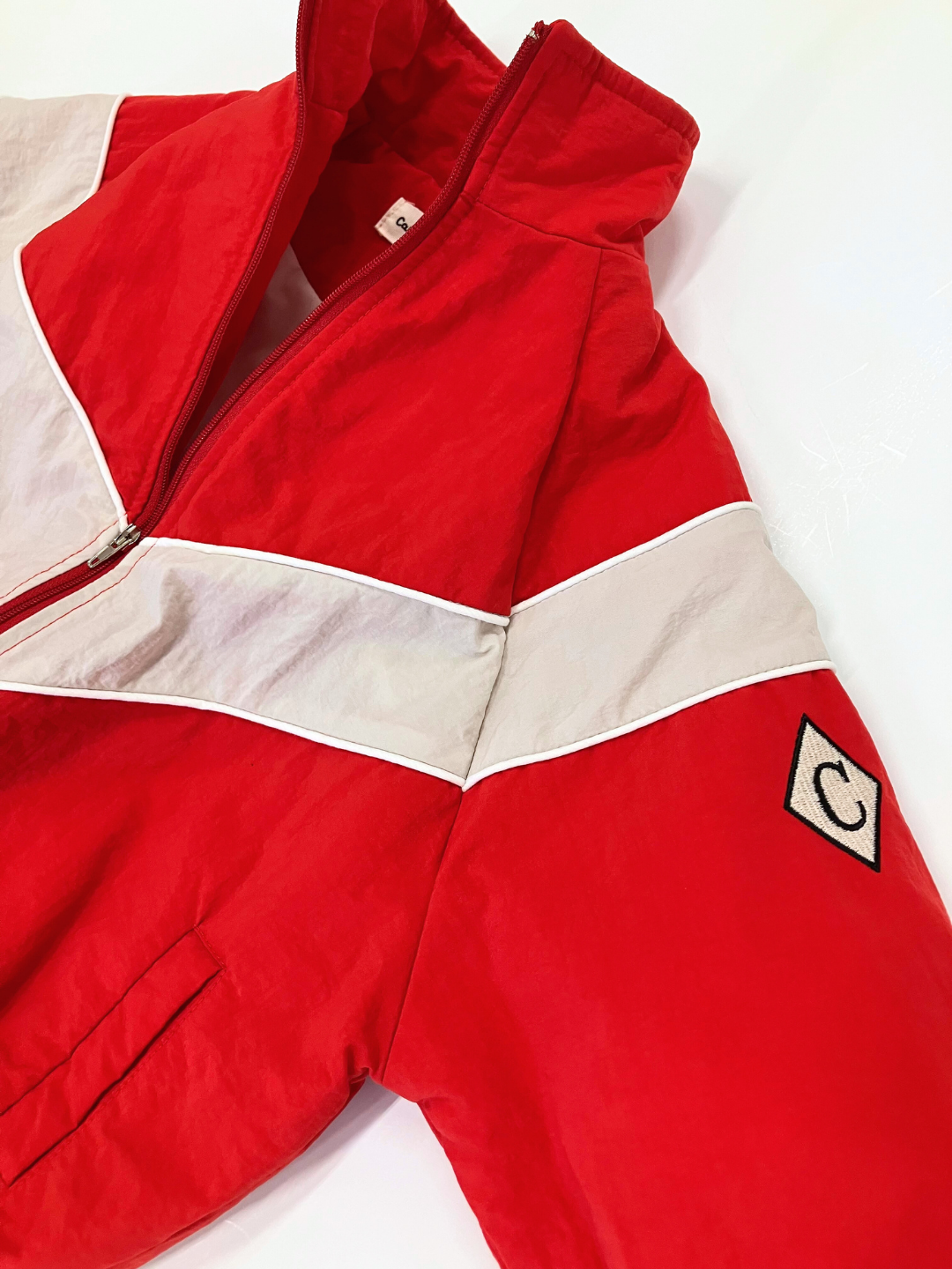 Red | A WARM-UP JACKET, designed as a lightweight layer by a renowned Korean label, is available in red and white with a zip-up front and features a retro design with the letter "C" logo on the sleeve.