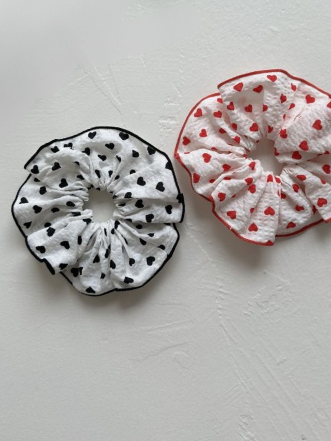 Two white hair scrunchies on a textured white surface, one is printed with small black hearts, and one with red hearts..