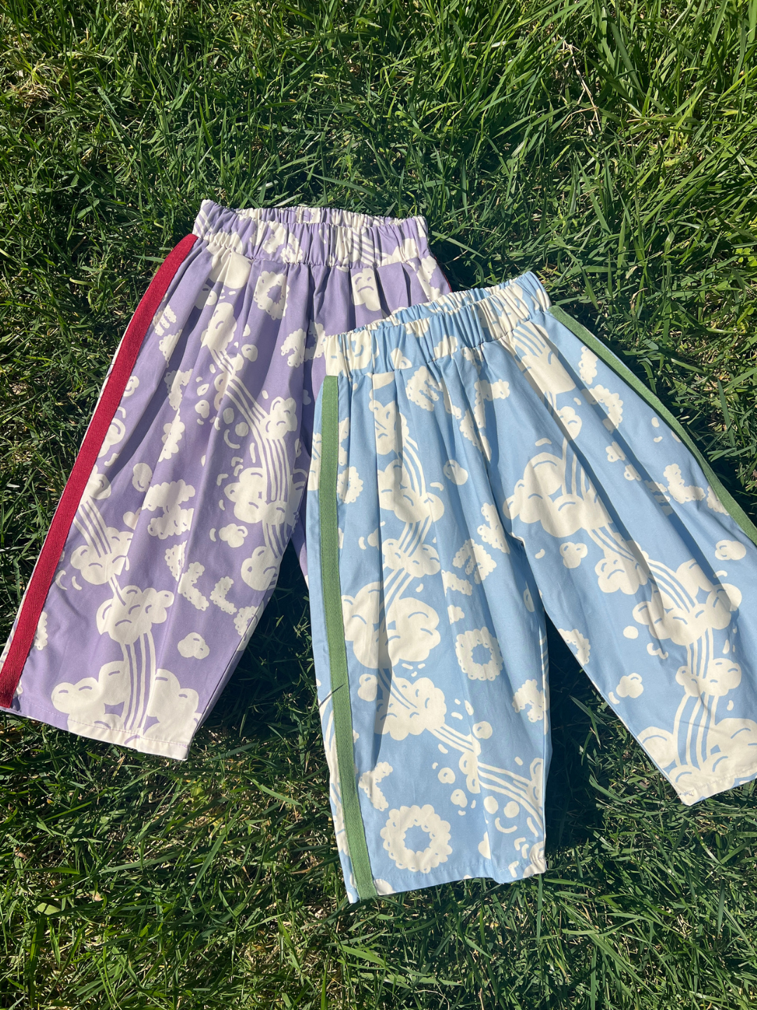 Blue | Two SKYWRITING PANT pairs with elastic waists, featuring cloud and rainbow patterns: one purple with a red side stripe and one blue with a green side stripe, shown lying on grass.