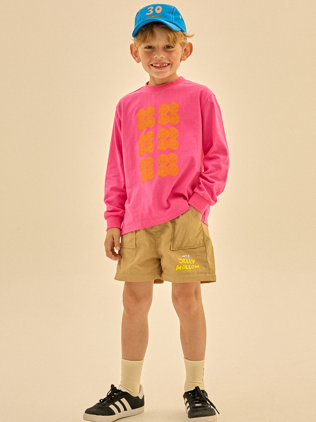 Child wearing Clover Long Sleeve T-shirt with a blue baseball hat, tan shorts, cream socks, and black shoes.