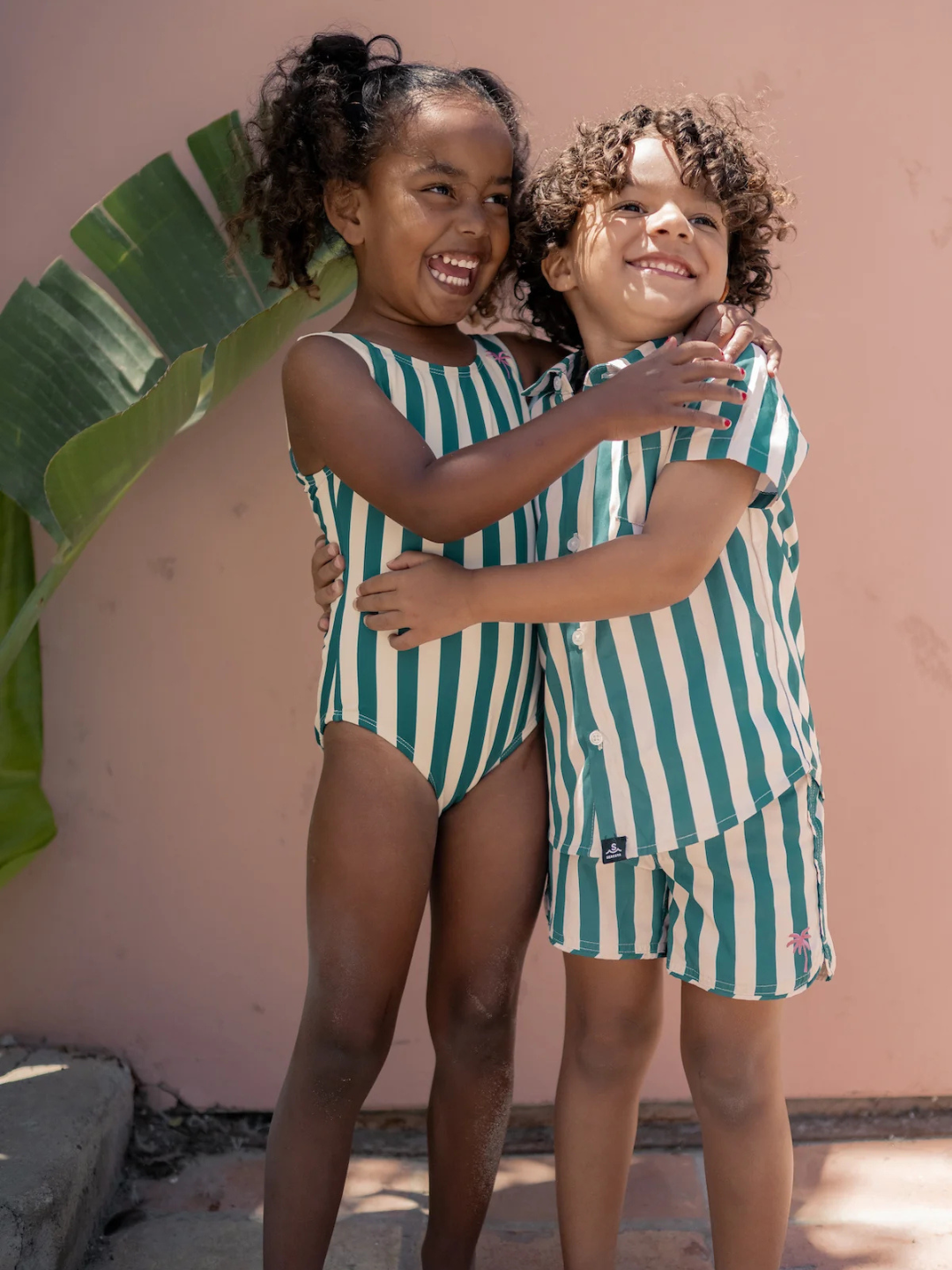 Two kids wearing CABANA STRIPE ONE PIECE SWIMSUITS made from sustainable fabric smile and hug each other outdoors by a pink wall and a large green leaf.