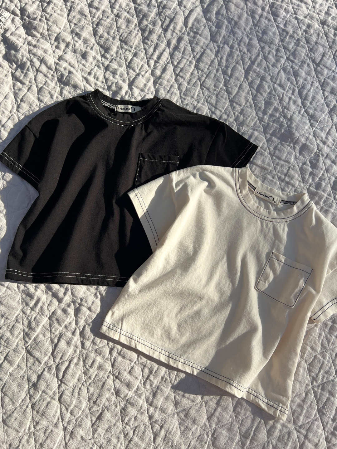 Black | Two of the kids' stitch tees are laid flat on a quilt in the sun