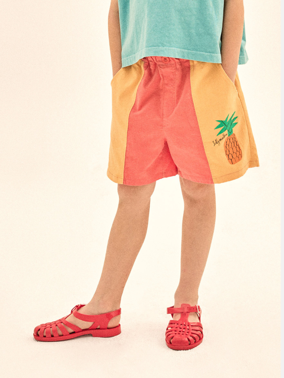 Child wearing colorful shorts with a pineapple design on a plain background, with red sandals.