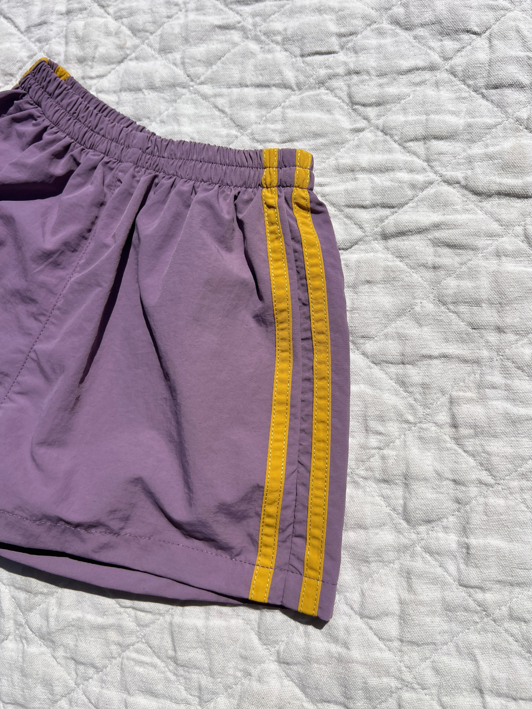 Purple | The SPRINTER SHORTS, featuring purple fabric, two yellow side stripes, and an elasticated waist, are displayed on a light quilted surface.