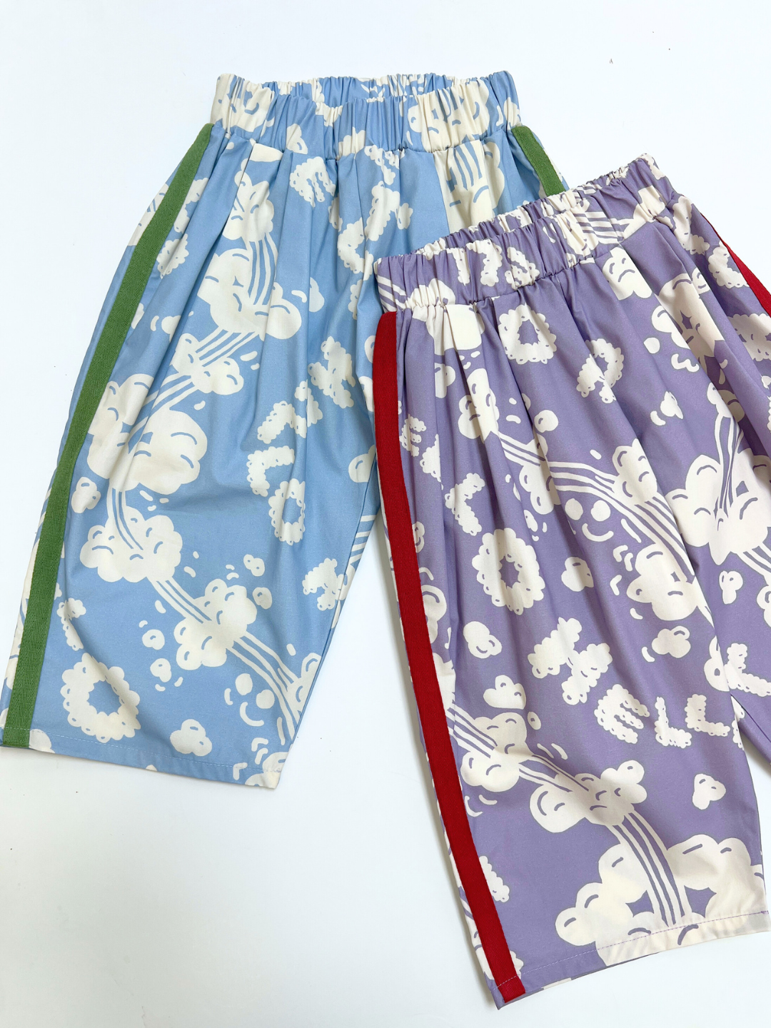 Two SKYWRITING PANT shorts with white floral patterns—one light blue with green side stripes and one light purple with red side stripes—displayed on a white background.