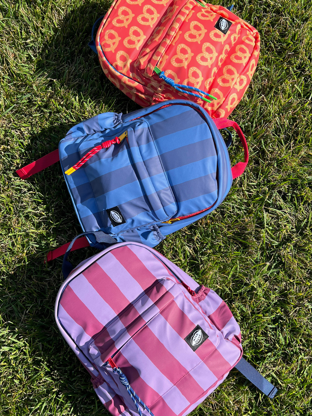 Three colorful kids backpacks from Milk Teeth are arranged diagonally on a green grass lawn. The top backpack is orange with a yellow pretzel pattern, the middle is blue with darker gray stripes, and the bottom is maroon and purple with vertical stripes.