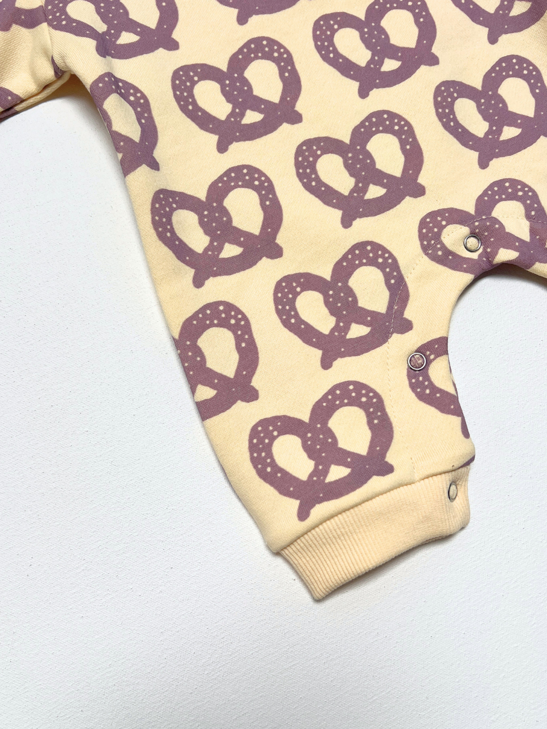 A close-up of a baby bodysuit featuring a repeating purple pretzel print on a warm cream background, with a ribbed ankle cuff and snap closures.