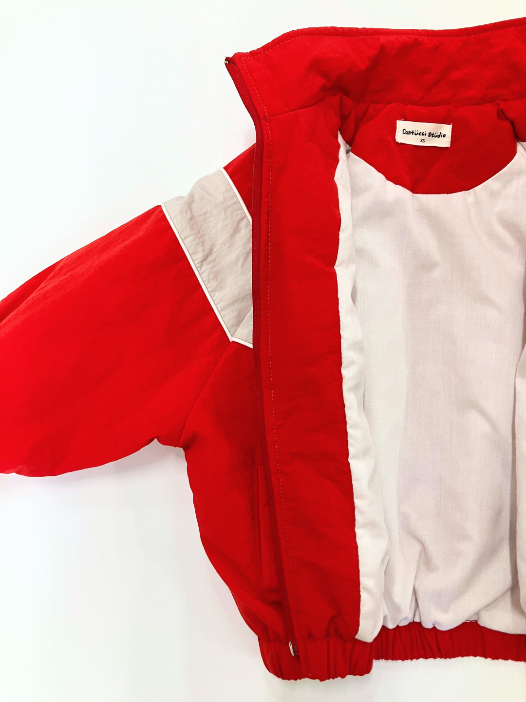 Red | A WARM-UP JACKET in red and white with a white lining, partially zipped open, displayed against a plain white background. This lightweight layer comes from the renowned Korean label "Cautious Studio.