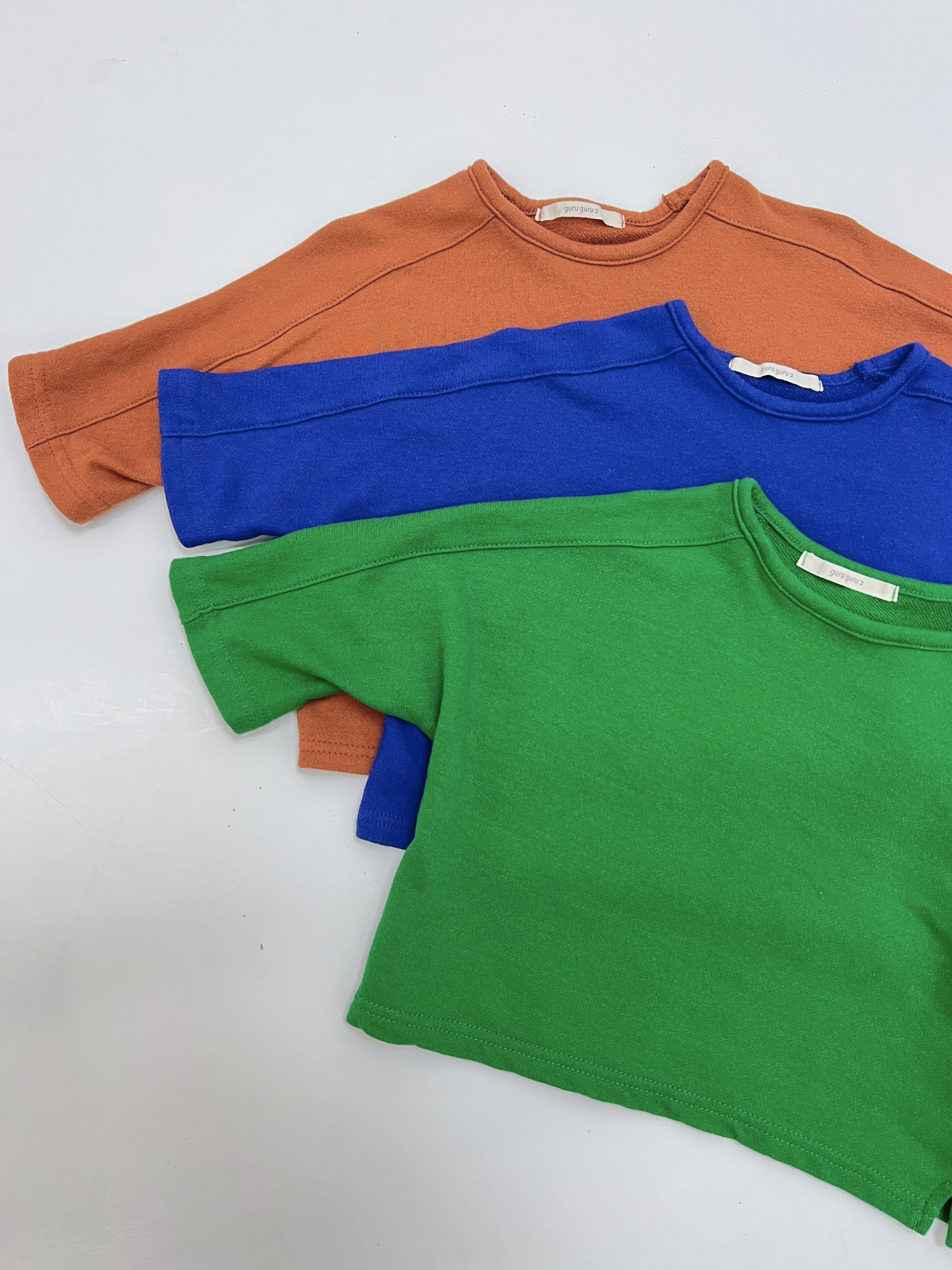 Three plain, long-sleeved BABY BOATNECK shirts in brown, blue, and green made from soft terry cotton are stacked on a white surface—a cozy essential from a trusted Korean baby brand.