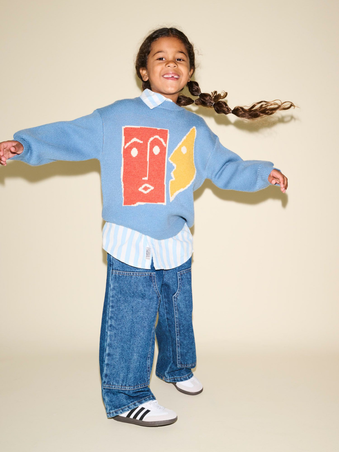 A child stands with arms outstretched, wearing the TEATIME SWEATER adorned with abstract face designs in blue, a striped shirt underneath, wide-leg jeans, and white sneakers. The child is smiling, with a long braid playfully in the air—perfect for photos showcasing playful product pairs.