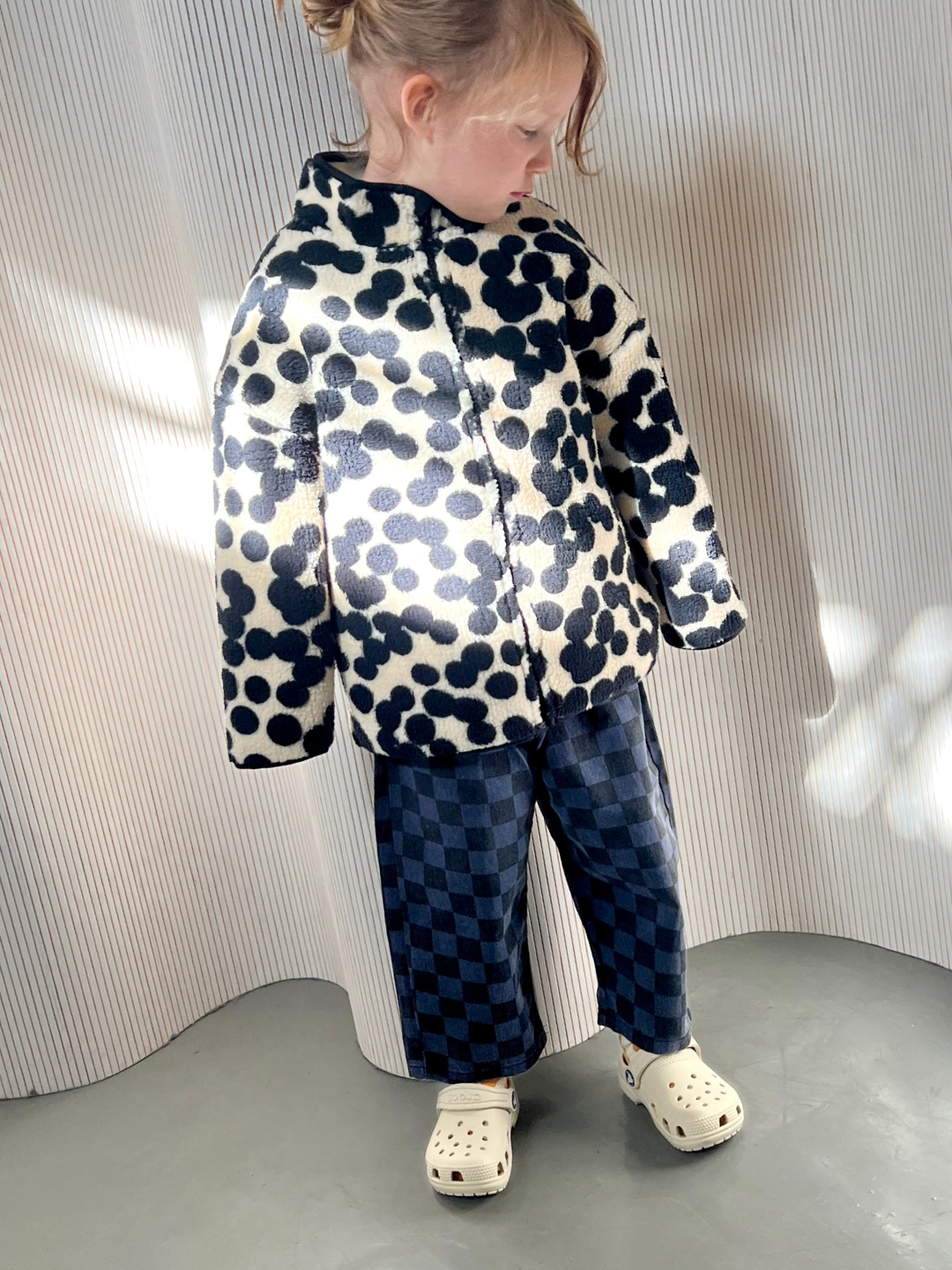 Blue | A child wearing the blue Casper pants in navy and black checkerboard, with a black and white spotted fleece jacket and white Crocs. They are standing on a grey floor in front of a wavy white wall.