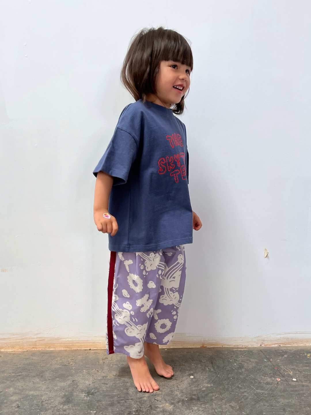 Purple | A young child stands barefoot indoors, wearing loose-fitting SKYWRITING PANT with a white floral pattern and a blue oversized T-shirt with red text.