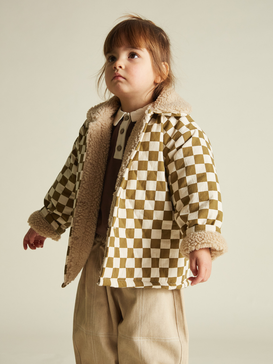 A young child stands slightly to the side, wearing the EMERY COCO CHECKERBOARD COAT—an oversized, brown and white unisex jacket with shearling trim—paired with beige pants and a brown shirt.