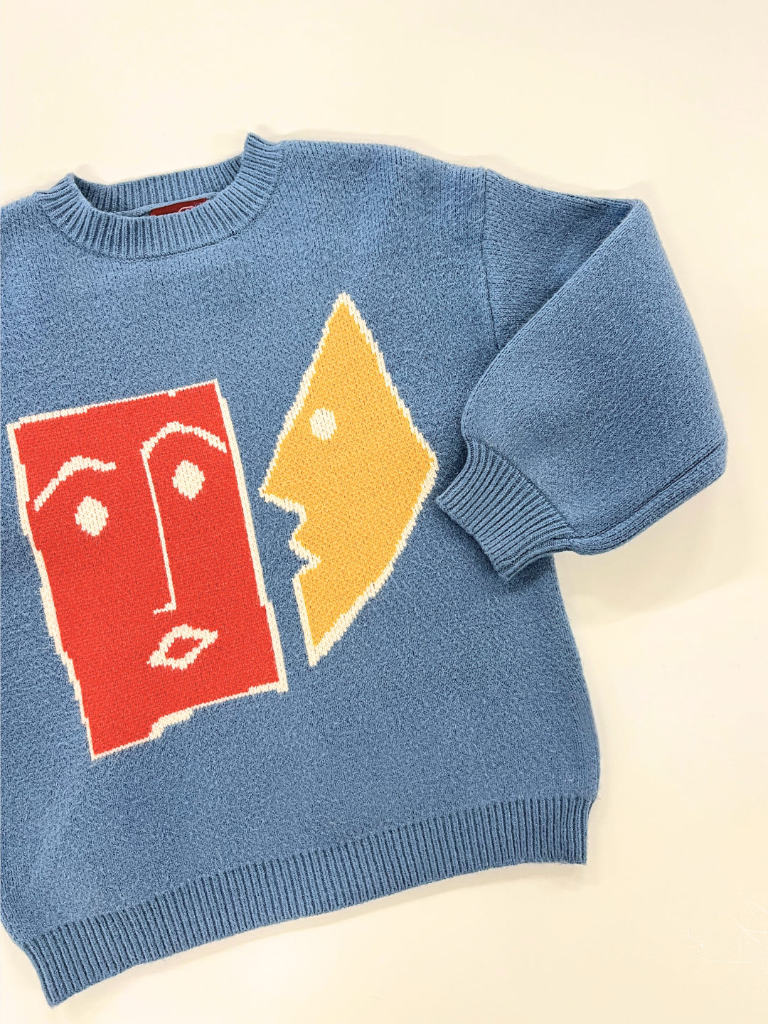 Slate | Introducing the TEATIME SWEATER, a stylish blue sweater featuring bold abstract face designs in vibrant red and yellow on the front. Displayed on a crisp white background, it's ideal for capturing stunning photos.