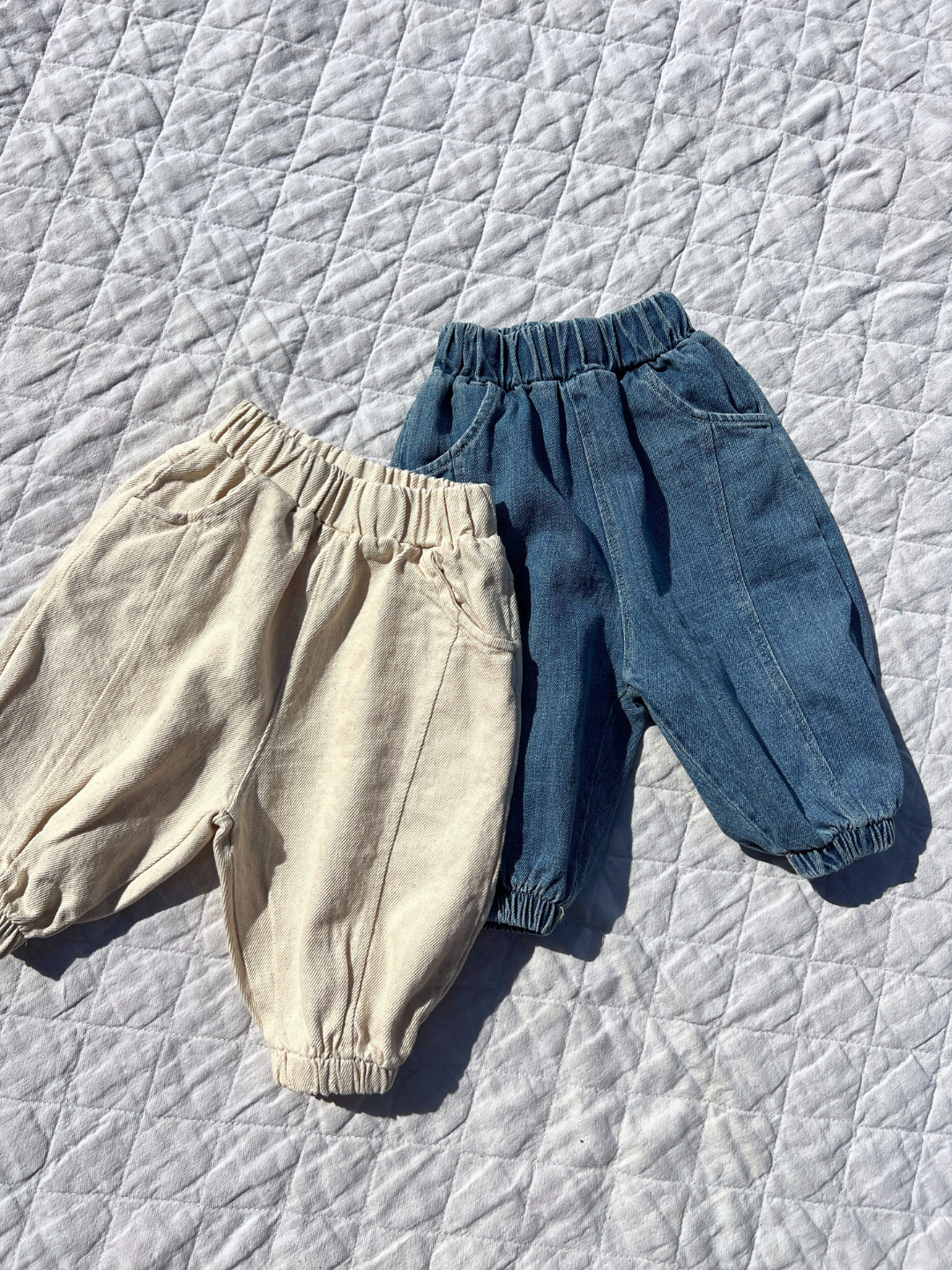 Ecru | Two pairs of toddler pants sit on a quilted surface, highlighting their jogger-style fit. One pair is light beige, and the other is blue denim. Both boast elastic waistbands and cuffs for active kids. Explore comfort with Valu Baby's BALLOON JEAN collection!.