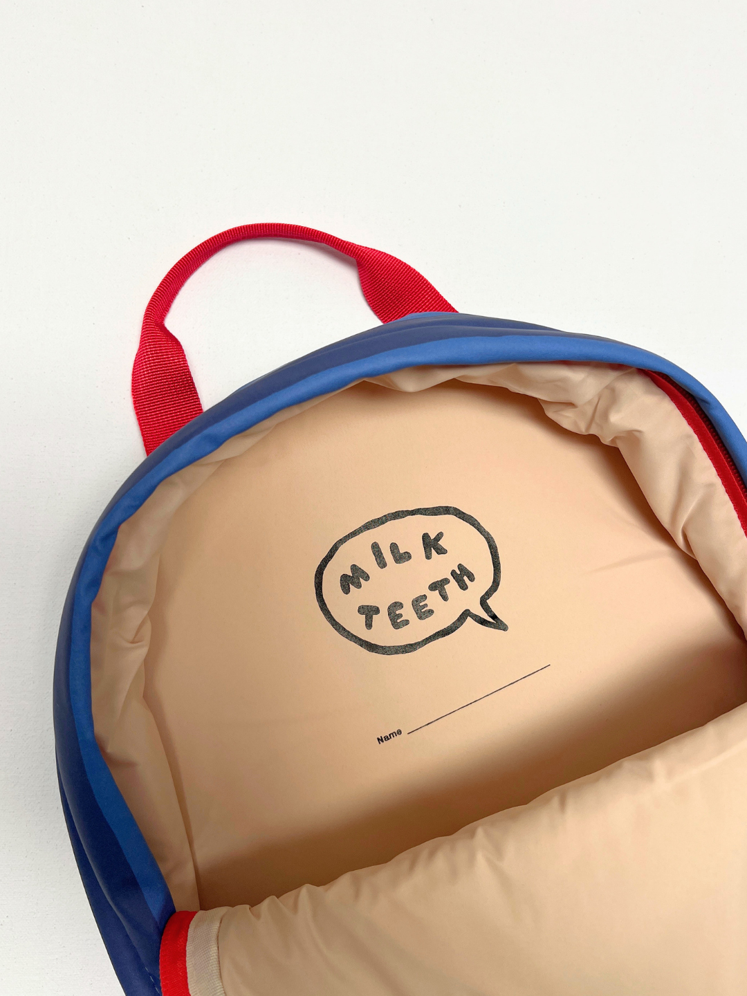 Close-up of the inside of a puffy quilted backpack with a beige interior, blue outer, red top handle, and a printed speech-bubble logo reading 'MILK TEETH' on the lining with a name label space.