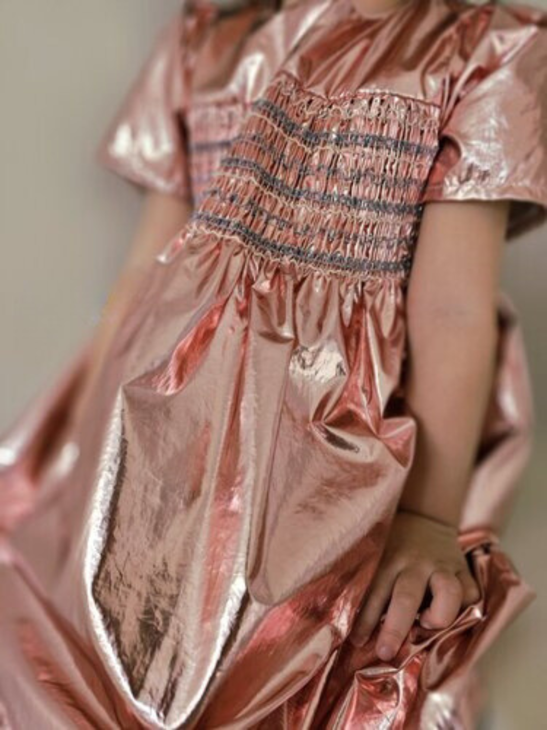 Cropped image of a child wearing a party dress in pink metallic fabric with a round neck, short sleeves, smocked bodice, empire waist and full skirt.