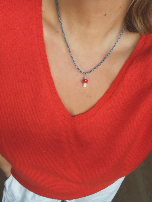 Second image of The FUNGI NECKLACE by Meyous jewelry is a handmade, genderless blue cord necklace with an adjustable fit and a red and white glass mushroom charm, shown on quilted white fabric—ideal for any style.
