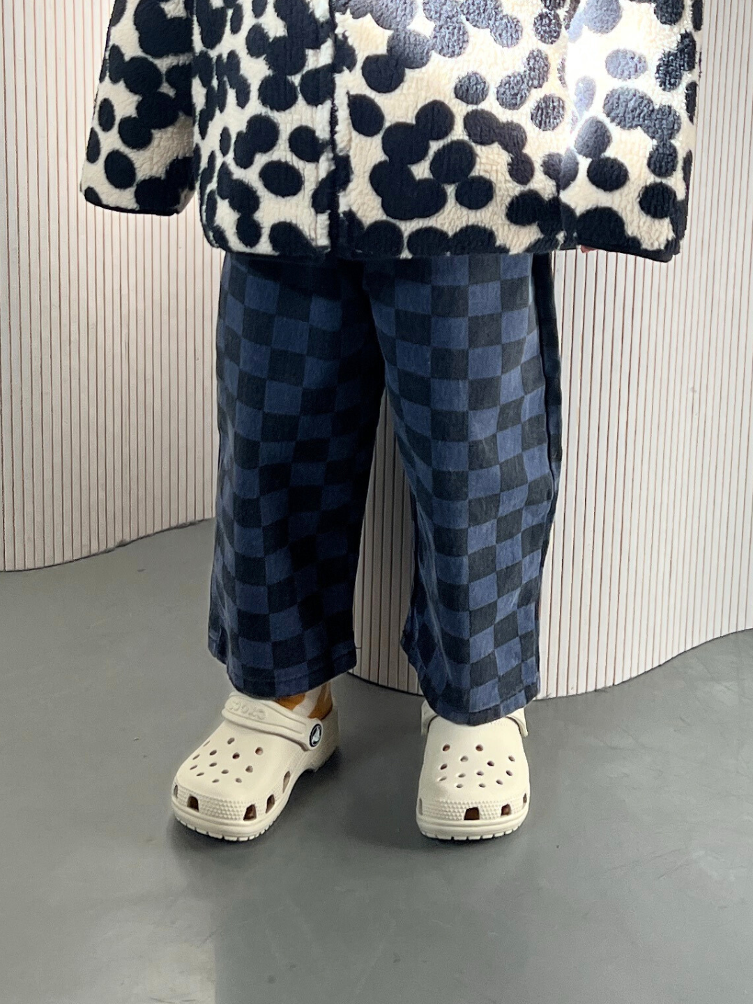 Blue | A child wearing the blue Casper pants in navy and black checkerboard, shown from the waist down. They are wearing a black and white spotted fleece jacket and white Crocs, and are standing on a grey floor in front of a wavy white wall.