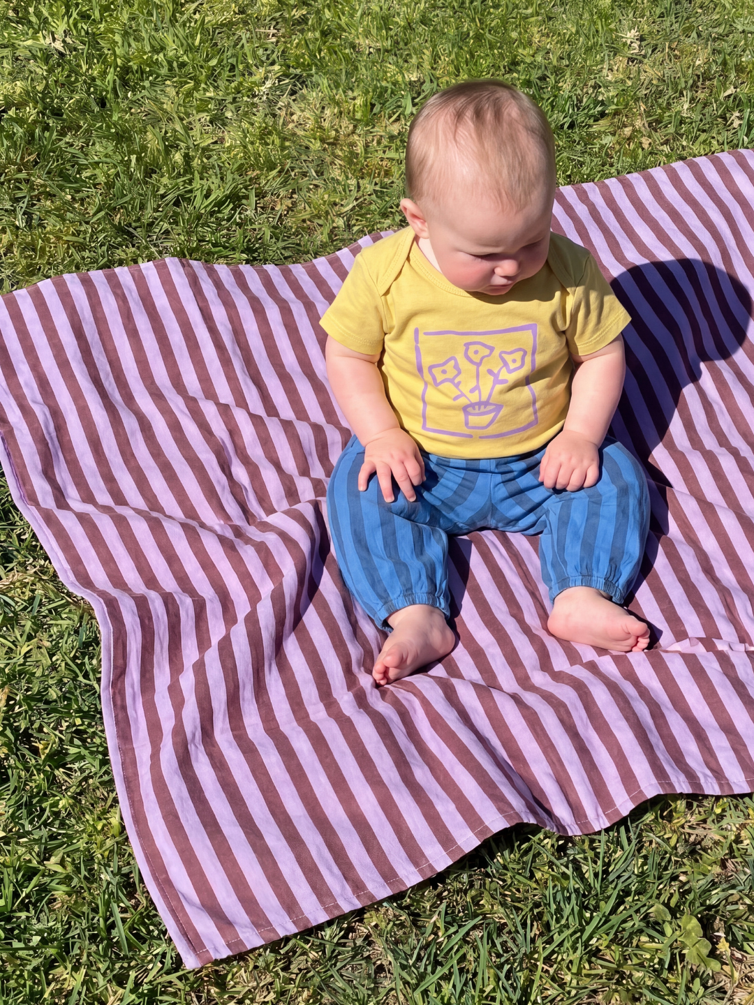 A baby wearing the striped organic cotton pants in blue stripes, with a yellow t-shirt with a flower motif, seated on a purple and maroon striped quilt, on grass.