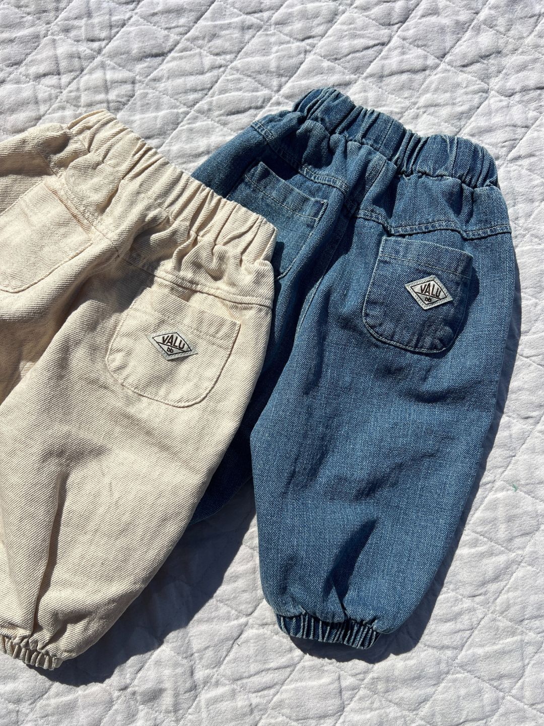 Ecru | Two pairs of BALLOON JEAN children's pants in jogger-style with elastic waists, one beige and one blue, featuring pockets with small fabric labels by Valu Baby, are displayed on a quilted surface.