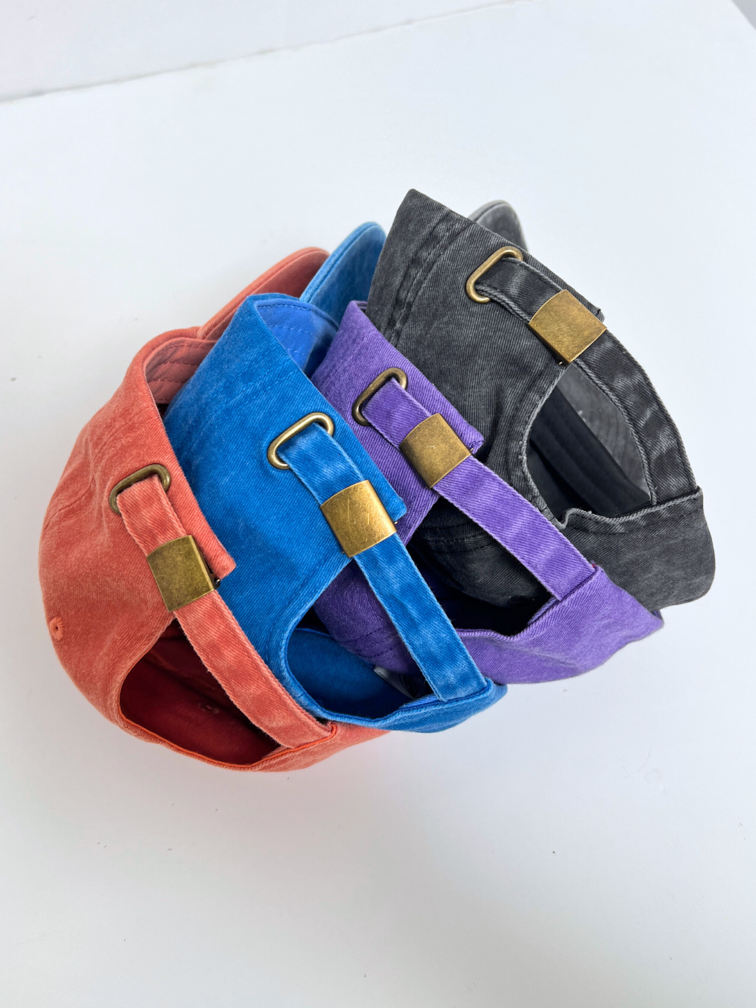 Four MT DAD CAPs in red, blue, purple, and black are stacked together, highlighting their adjustable brass buckle closures. Made from durable cotton canvas, these caps stand out against a white background.
