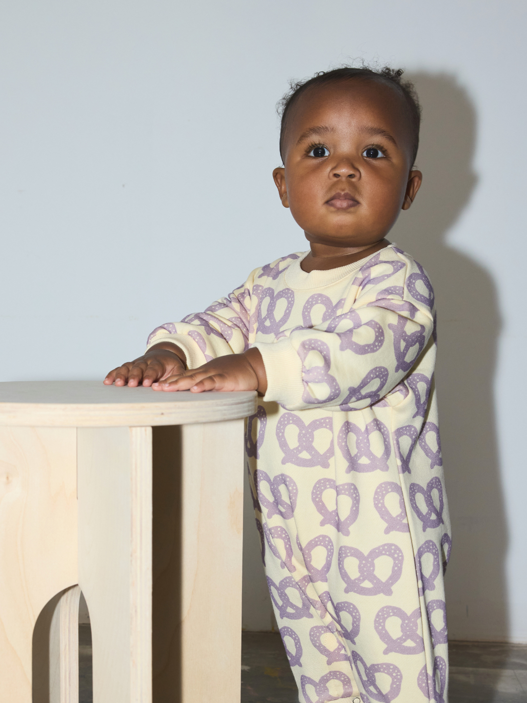 Purple/Yellow | A baby wearing a cream-colored long-sleeve footless onesie with a purple pretzel pattern, standing next to a light wood stool against a plain wall.