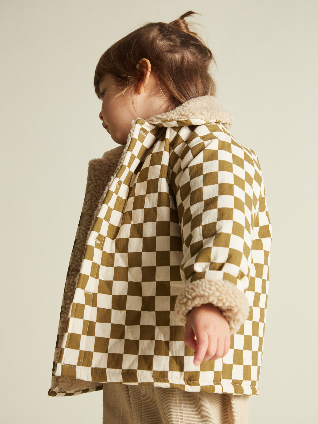 A young child stands sideways against a plain background, wearing the EMERY COCO CHECKERBOARD COAT with a shearling collar and cuffs, paired with light-colored pants.