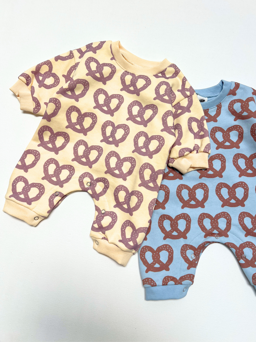 Two baby long-sleeve footless onesies laid flat on a white background. The front one is cream with a repeating brown pretzel pattern, and the one behind it is light blue with a repeating brown pretzel pattern.