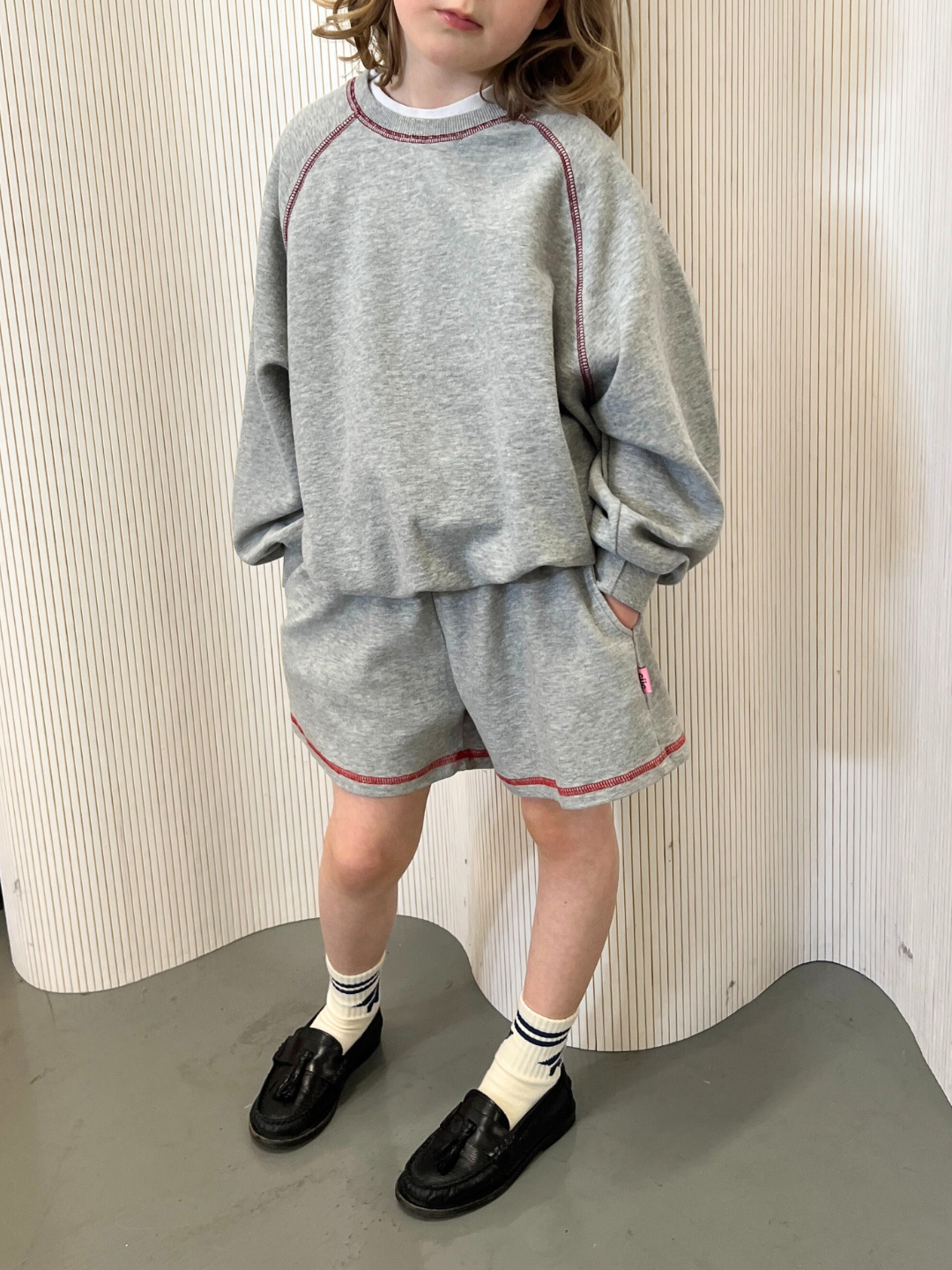 Grey | A child in a Cantucci Studios ensemble stands against a striped background on a grey floor, wearing the VARSITY SWEATSHIRT, retro-styled and made from brushed cotton, paired with grey shorts featuring red stitching, black loafers, and white socks.