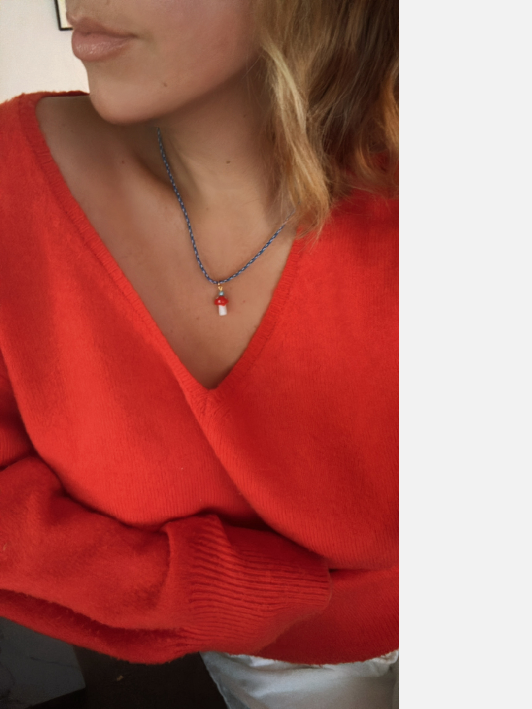 A person wears a red V-neck sweater and the FUNGI NECKLACE—a blue beaded necklace with a small red-and-white glass mushroom charm from Meyous jewelry. Only their lower face and upper torso are visible.