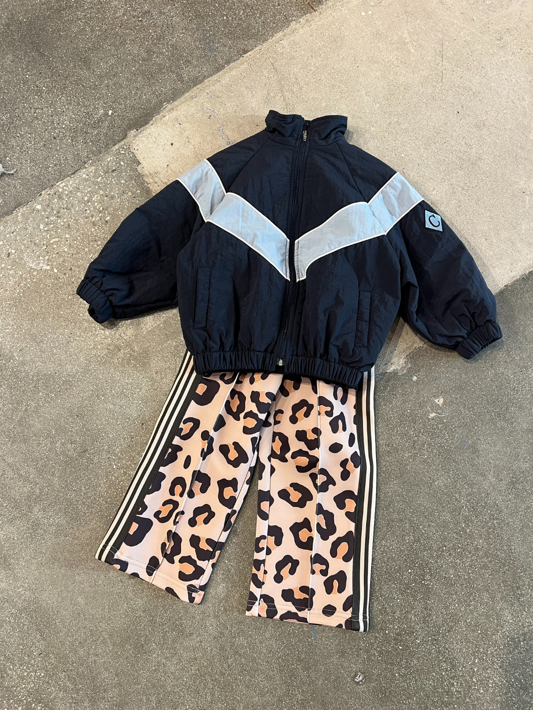 Navy | Child's outfit laid out: the WARM-UP JACKET in black and gray with a chevron stripe, paired with pink leopard print pants featuring black and white side stripes.