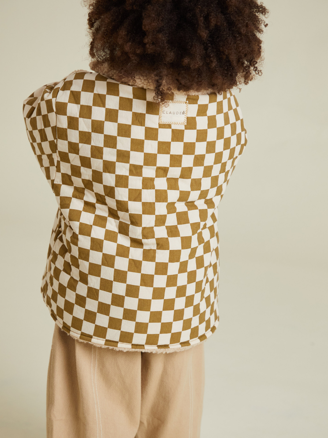 A child with curly hair stands with their back to the camera, wearing the EMERY COCO CHECKERBOARD COAT and beige pants against a neutral background.