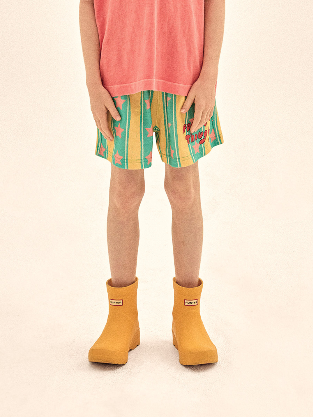 Child wearing colorful striped shorts and mustard yellow rain boots on a beige background