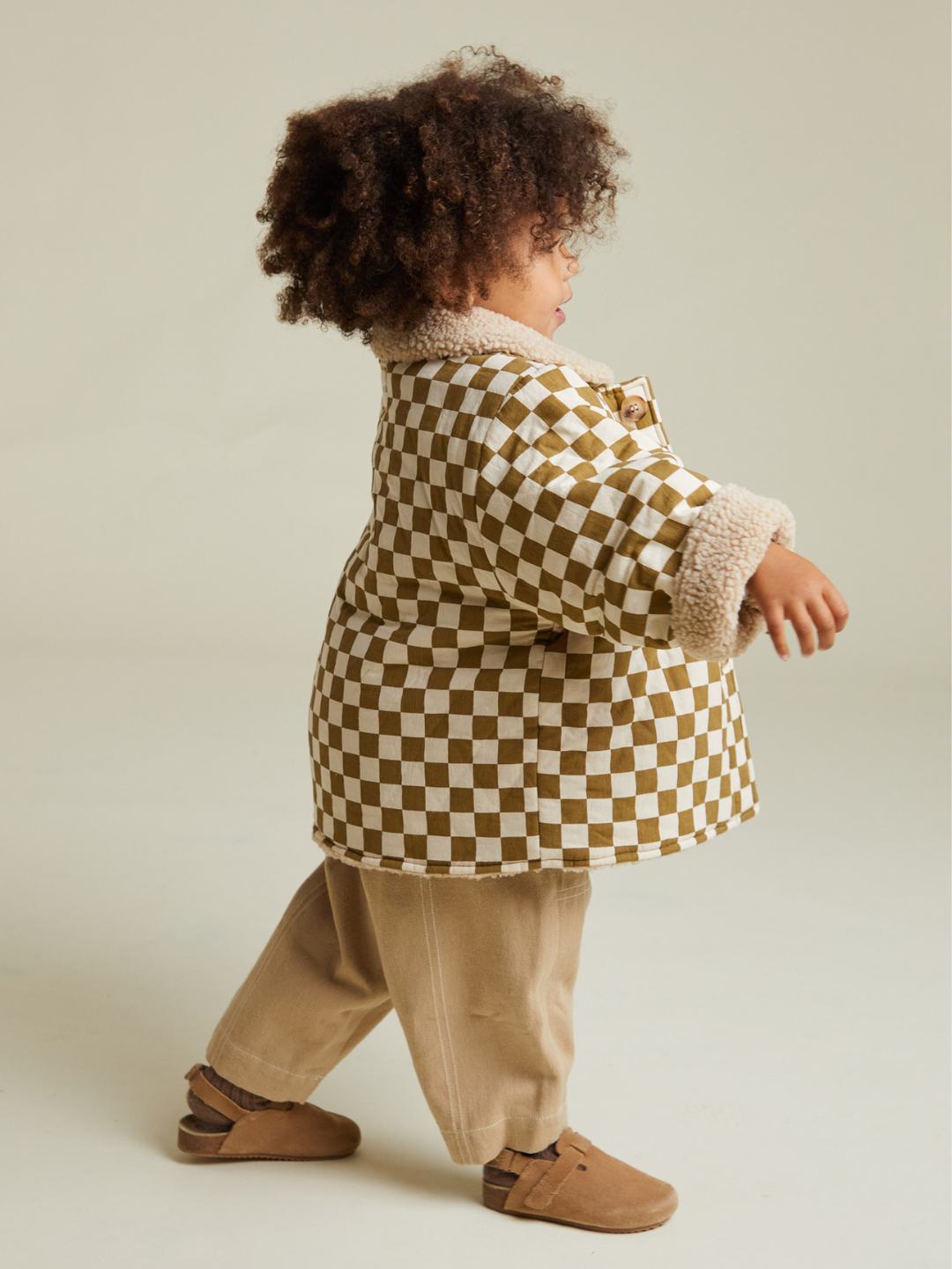 A young child wearing the EMERY COCO CHECKERBOARD COAT, beige pants, and brown shoes walks against a plain light background.