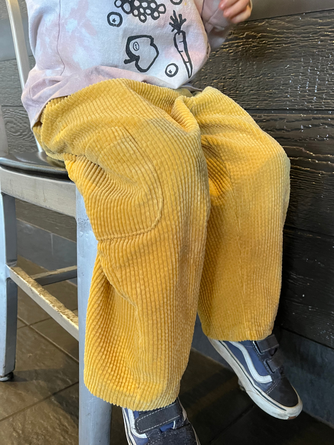 Gold | A child, dressed in a pink sweater adorned with cartoon designs and wearing comfy yellow CORD POCKET PANT from a trendy Korean kids brand, sits comfortably on a chair.