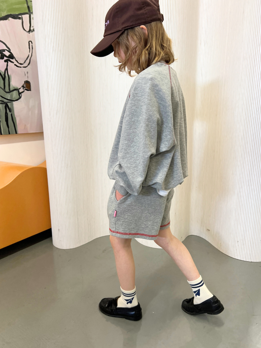 A child walks inside in a retro-style Cantucci Studios set, featuring the VARSITY SWEATSHIRT and shorts. With black shoes, white socks, and a dark cap, they tread on a gray floor against a textured white backdrop, exuding timeless fashion elegance.