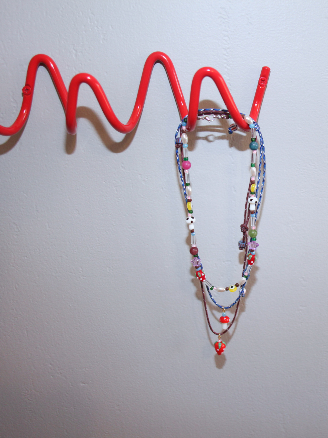 The FUNGI NECKLACE, featuring multicolored beads and a glass mushroom charm, hangs from a red wavy wall hook against a plain light gray wall.