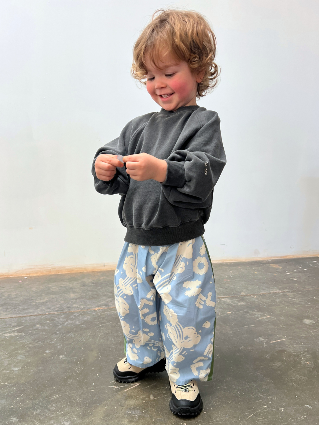A young child with curly hair smiles indoors, looking down while wearing a dark sweatshirt, black-and-white shoes, and wide blue floral SKYWRITING PANT.
