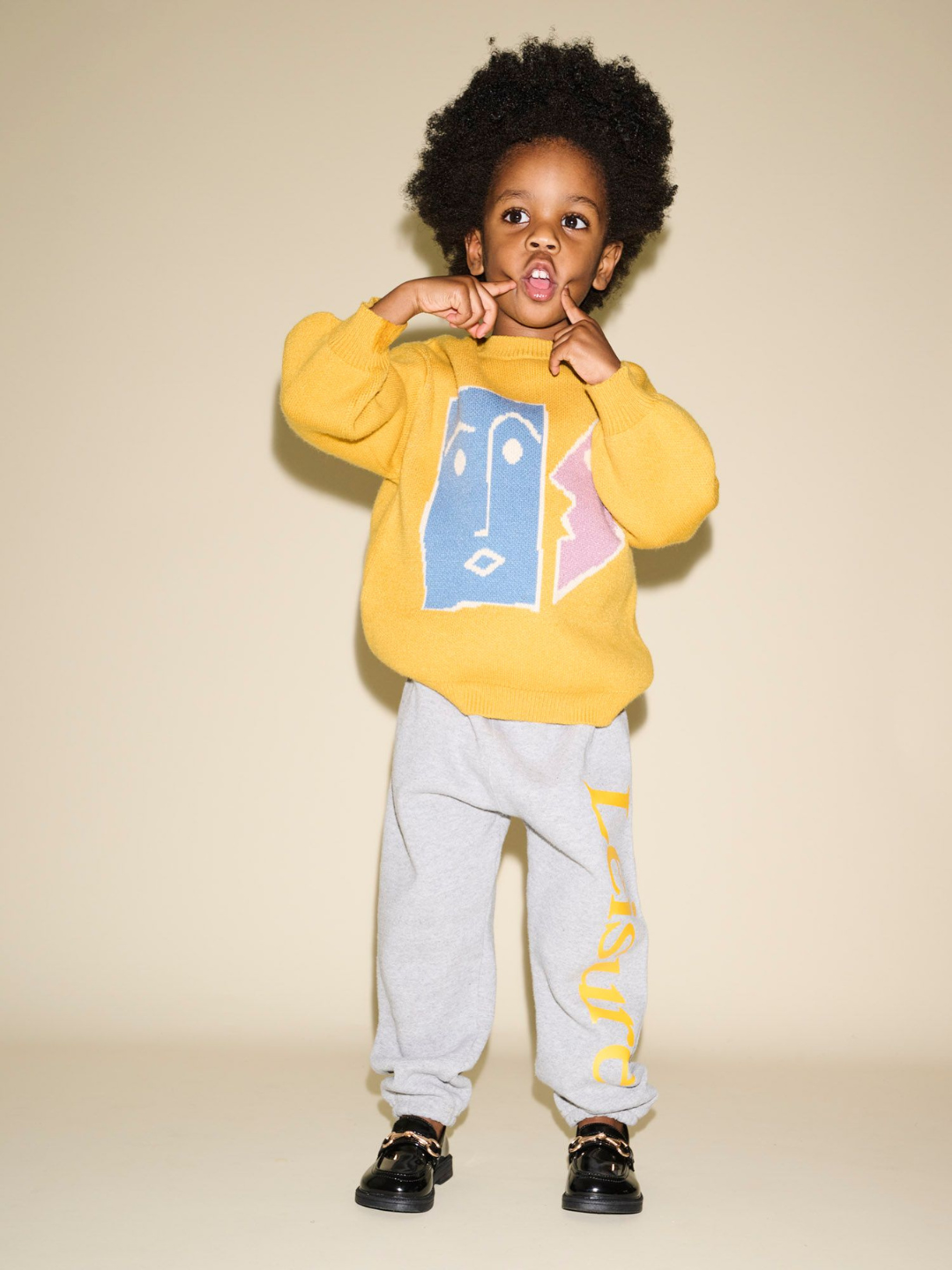 A young child stands against a neutral background wearing the TEATIME SWEATER in yellow, featuring a face graphic, and grey sweatpants with "Lacug" written on the left leg, hands raised near their face. Perfect for photos, this outfit makes for delightful product pairs in any kids' wardrobe.