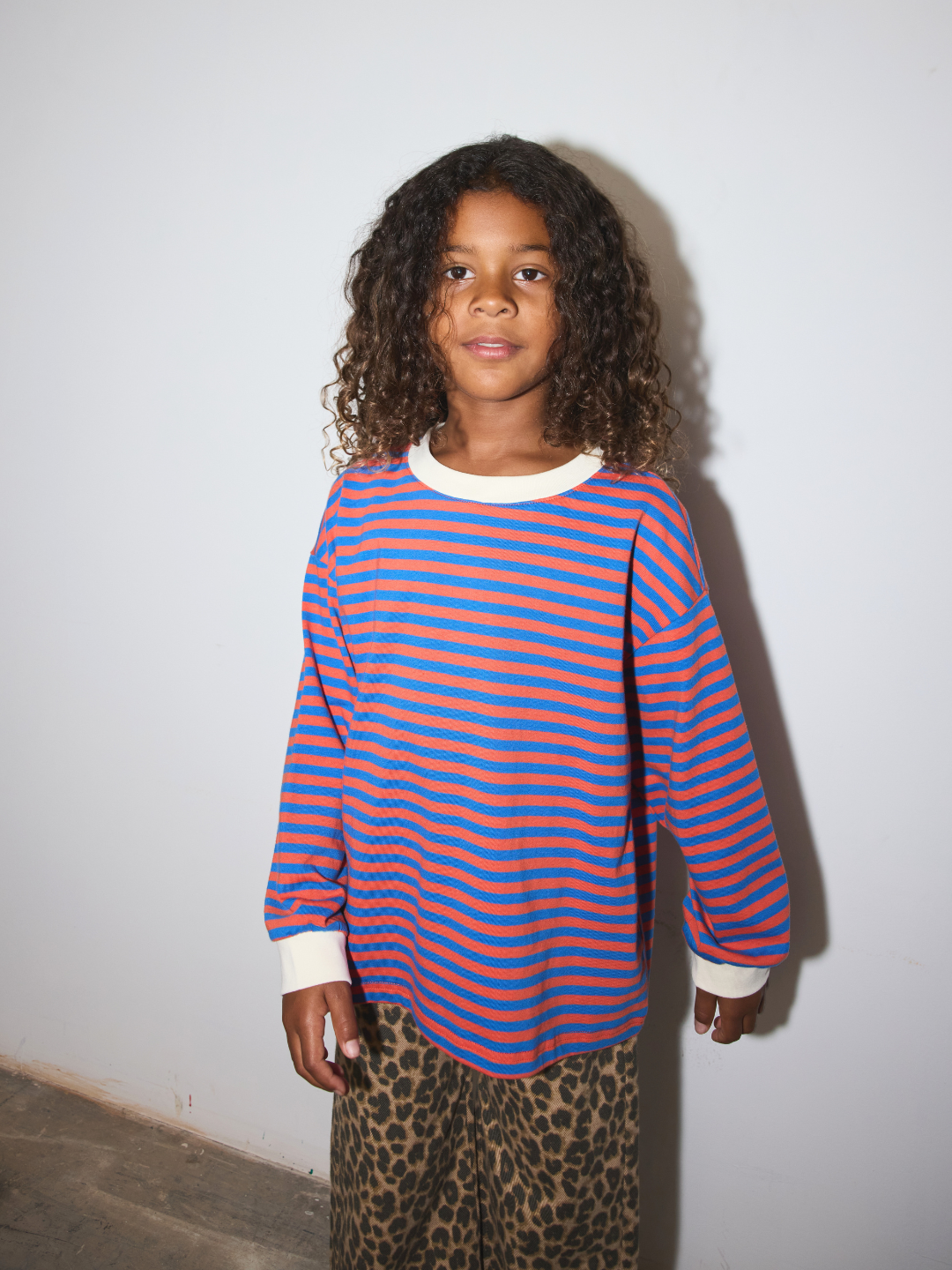 Red/Blue | A child wearing a long-sleeve crewneck shirt with horizontal blue and orange stripes, paired with leopard print pants, standing against a white wall.